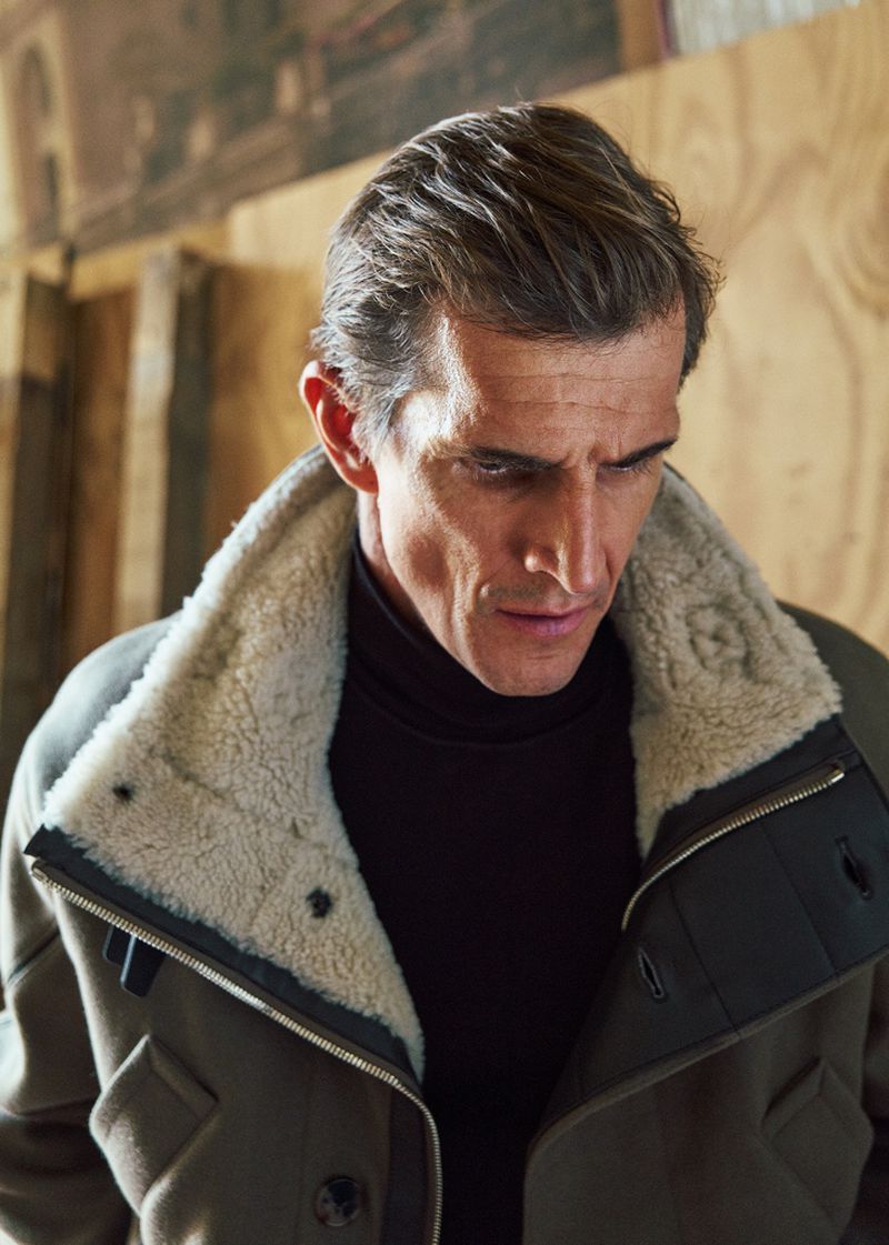Portrait of a mature man wearing a heavy shearling-collared coat and black turtleneck in an indoor wooden workshop setting.