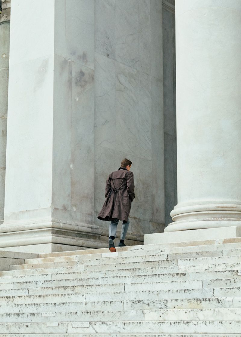 Man wearing a brown coat walking up large marble stairs between monumental neoclassical pillars.