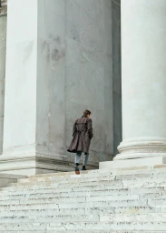 Man wearing a brown coat walking up large marble stairs between monumental neoclassical pillars.