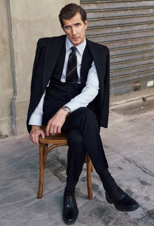 A man in a black suit, vest, and tie sitting on a wooden chair outdoors against a concrete wall and metal shutter.