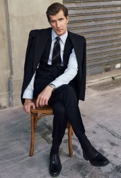 A man in a black suit, vest, and tie sitting on a wooden chair outdoors against a concrete wall and metal shutter.