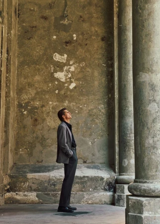 A man in a grey blazer and dark trousers stands profile against a weathered classical architectural backdrop with columns.