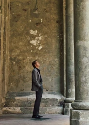A man in a grey blazer and dark trousers stands profile against a weathered classical architectural backdrop with columns.
