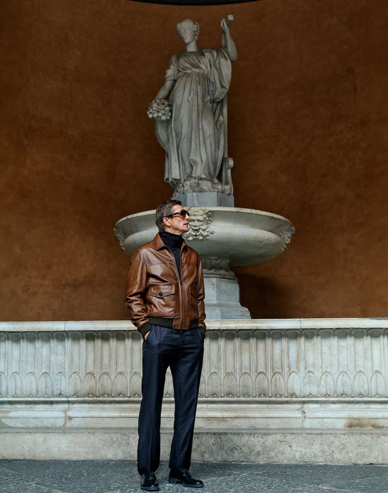 Man wearing a brown leather jacket and sunglasses standing in front of a stone fountain with a classical statue.