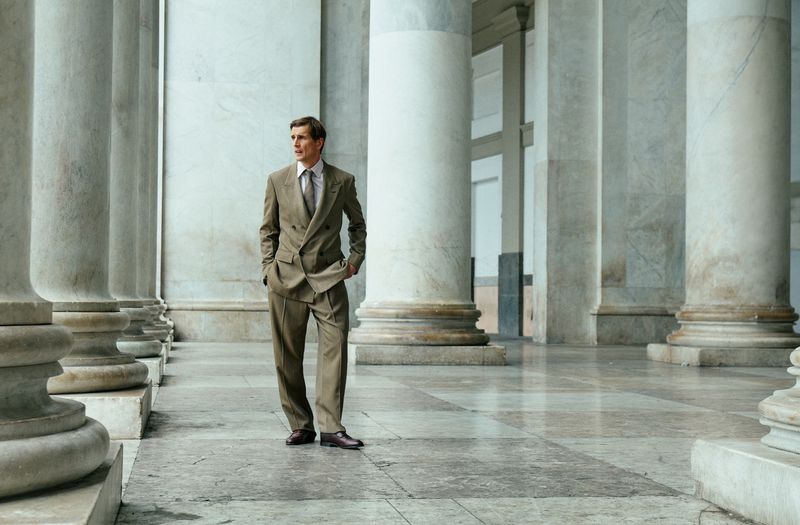 Man standing in a tailored taupe suit posing among large neoclassical marble columns in a grand hall