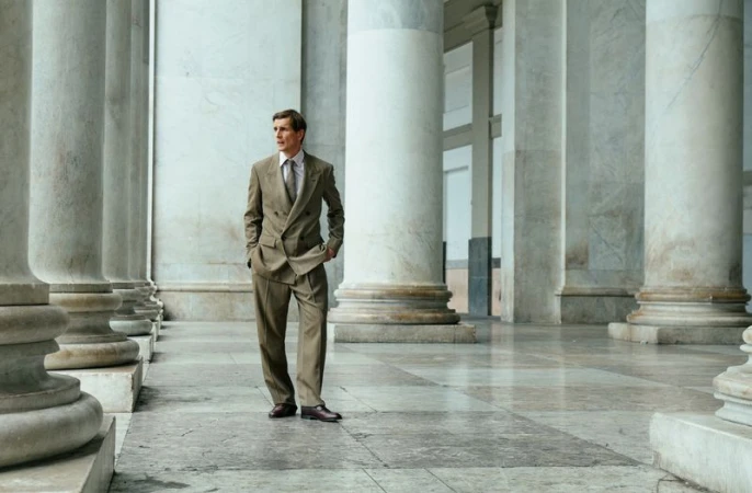 Man standing in a tailored taupe suit posing among large neoclassical marble columns in a grand hall