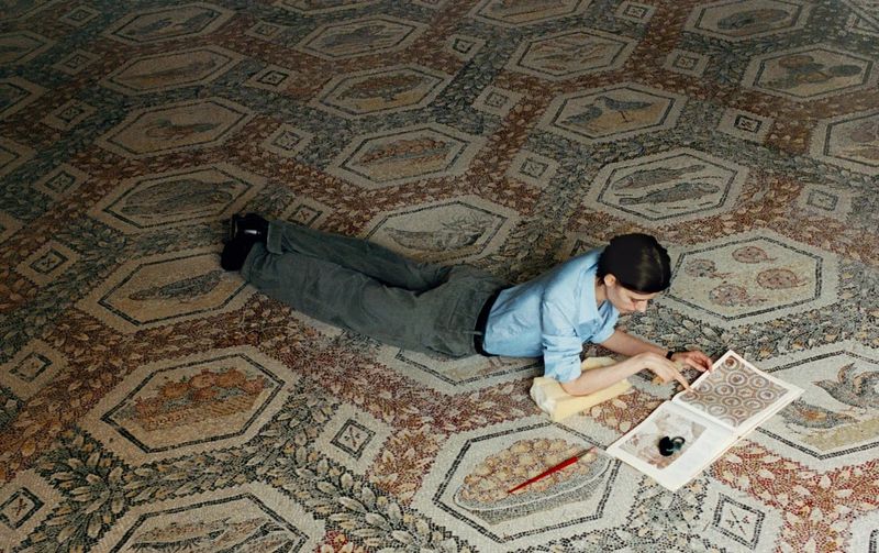 Model lying on stomach examining an open illustrated book spread out on an intricate ancient mosaic floor.
