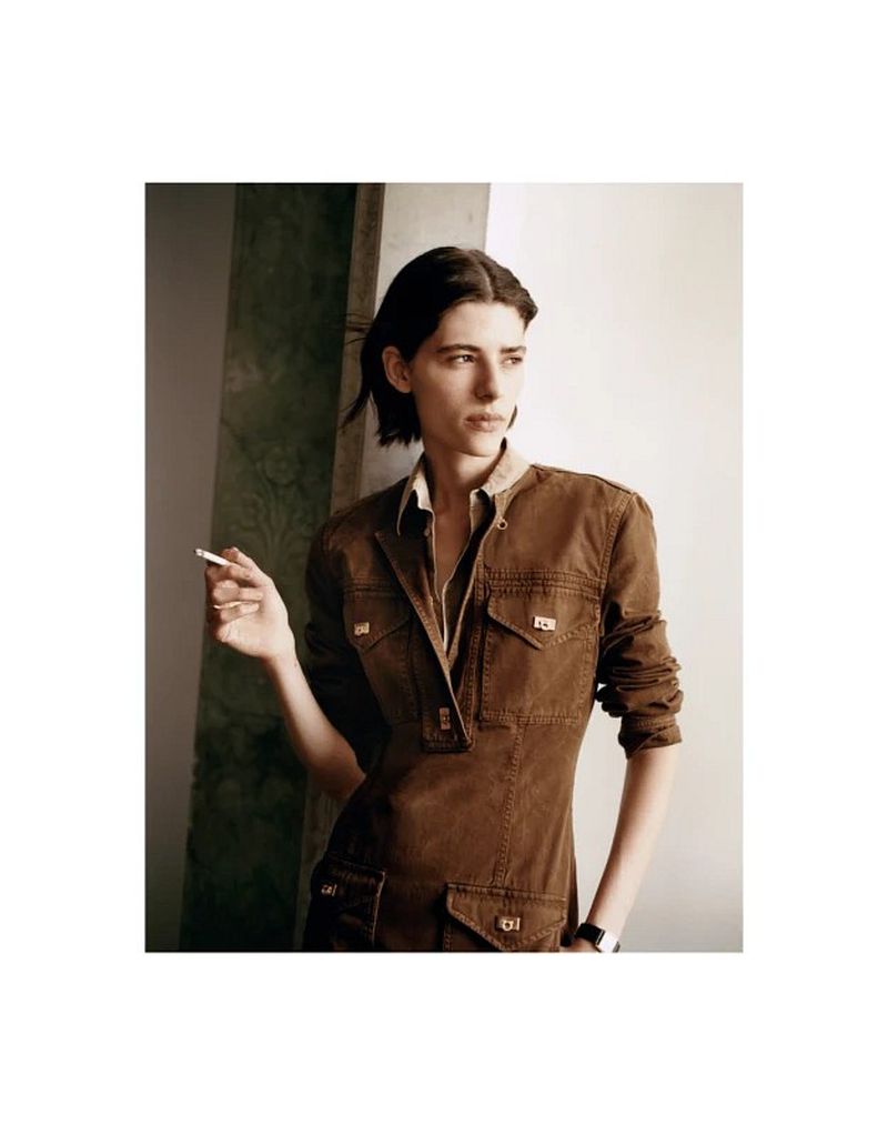 Model with dark hair wearing a brown utility jacket holding a cigarette against an architectural background