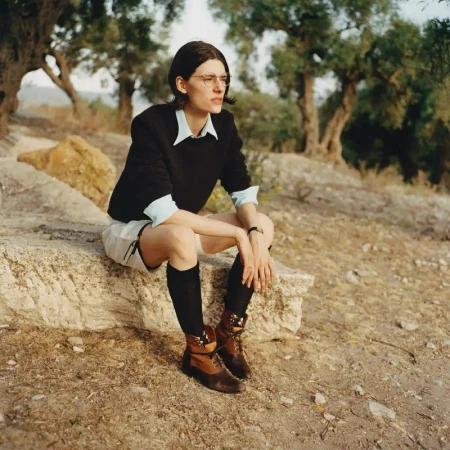 Model wearing dark sweater, light shirt, shorts, knee-high socks, and boots sitting outdoors on a stone structure near trees.