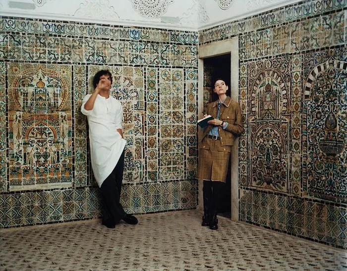A man in a white tunic and a woman in a plaid jacket and skirt lean against ornate tiled walls in a traditional interior