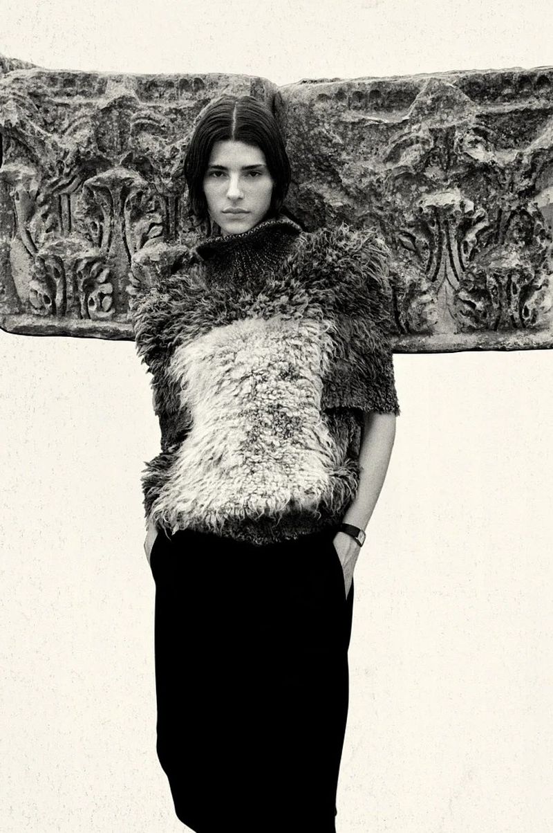 Model wearing a textured wool top and dark trousers posing against an ancient carved stone architectural block.