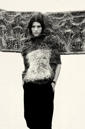 Model wearing a textured wool top and dark trousers posing against an ancient carved stone architectural block.