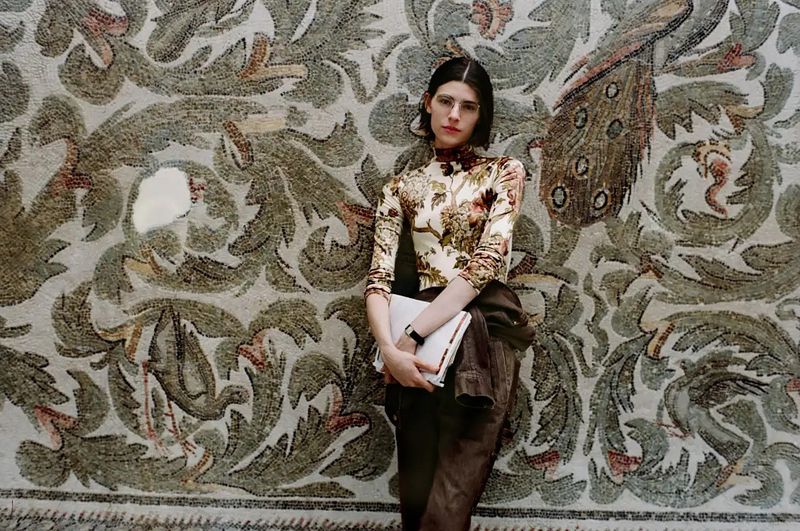 Model standing against an ornate antique mosaic wall holding a white folder, photographed by Brett Lloyd for Sotheby's.