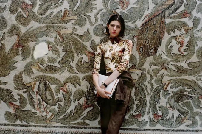 Model standing against an ornate antique mosaic wall holding a white folder, photographed by Brett Lloyd for Sotheby's.