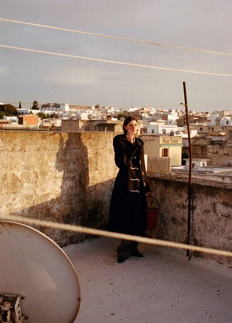 A woman in a dark coat standing on a sunny rooftop overlooking a town with distant buildings and warm light.