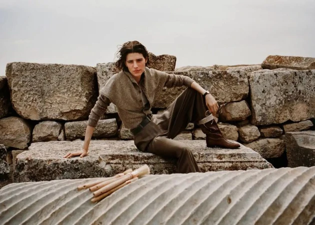 Model seated on ancient stone ruins wearing a brown knit top and dark trousers, photographed outdoors under an overcast sky.