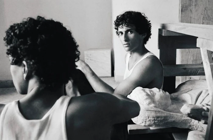 Monochrome image of two men in white tank tops working near a sculpted stone bust in an interior space