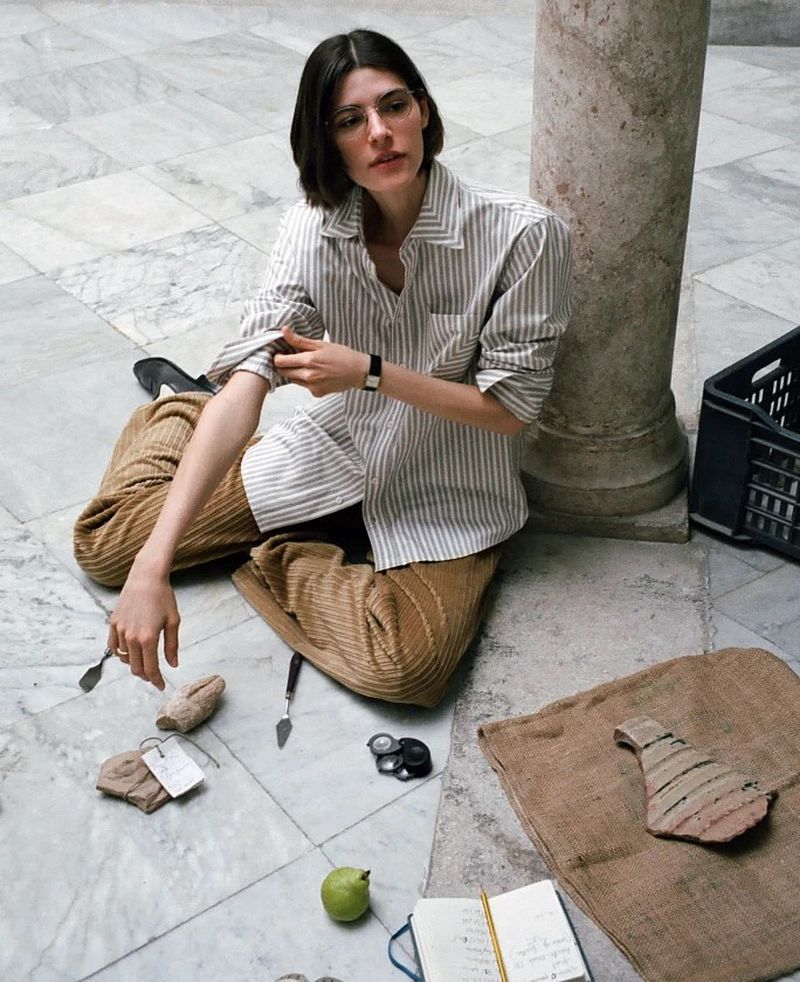 Model with dark hair rolling up the sleeve of a striped shirt while seated on a marble floor amidst artifacts.