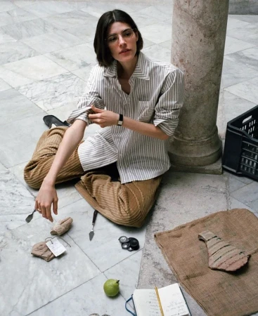 Model with dark hair rolling up the sleeve of a striped shirt while seated on a marble floor amidst artifacts.