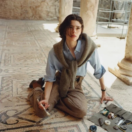 Model seated on an ornate mosaic floor wearing brown trousers, a light blue button-down shirt, and a brown sweater draped over shoulders.
