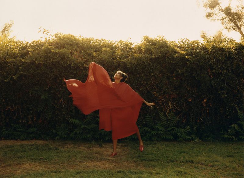 Woman in a flowing red dress dancing outdoors with fabric billowing against a dark green hedge background.