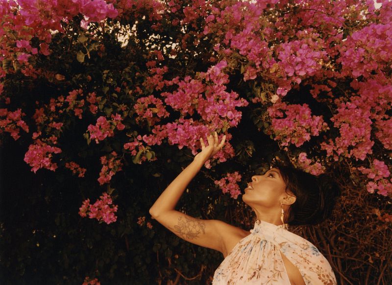 Woman with an arm tattoo reaching up toward vibrant pink bougainvillea flowers in a portrait for Cero Magazine.