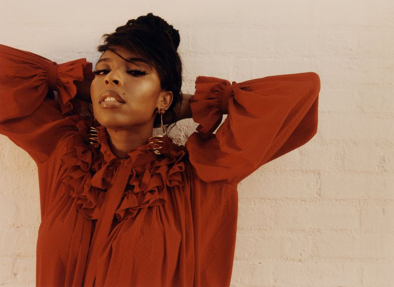 Portrait of Jessica Williams in a rust red ruffled blouse with statement gold earrings against a light textured wall.