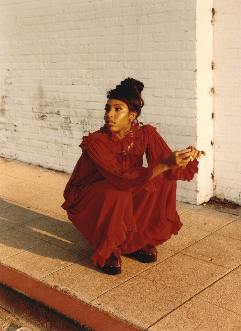 Black woman wearing a long red dress crouched against a light-coloured brick wall in warm evening sunlight.