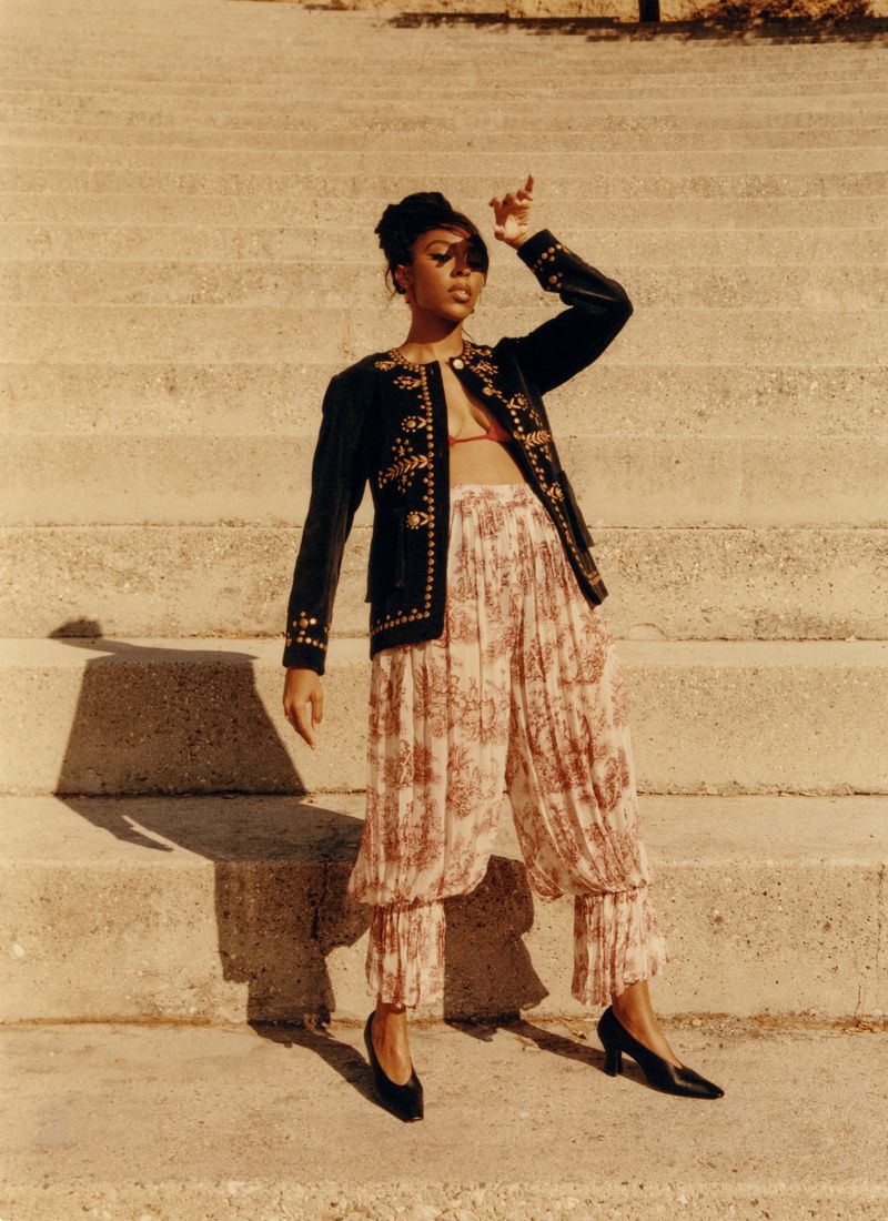 Woman standing on concrete steps wearing a dark embroidered jacket and patterned trousers for Cero Magazine editorial
