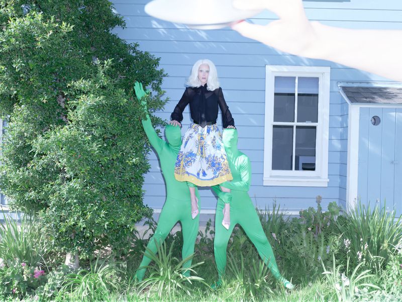 Model with white hair wearing a black blouse and patterned skirt lifted by two people in green bodysuits outside a blue house