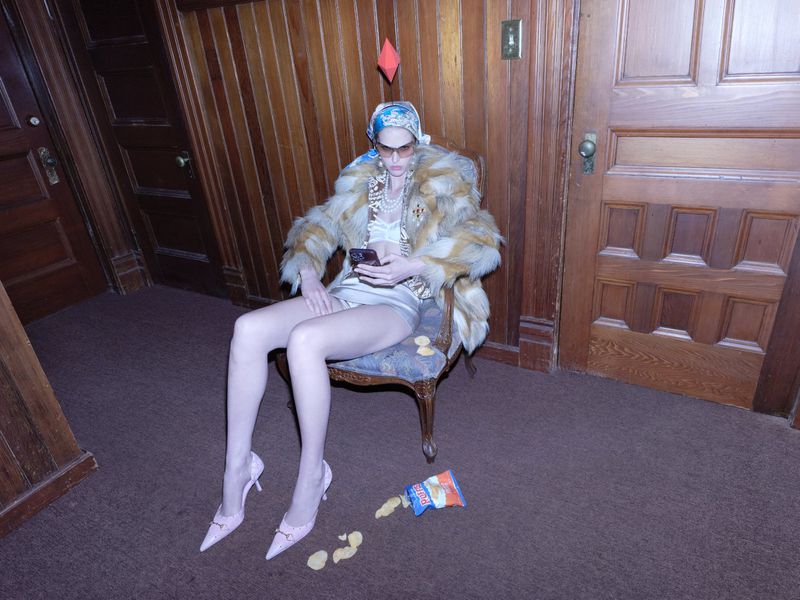 Model Abby Champion seated in a wooden chair holding a phone with a simulated red plumbob floating overhead in a wood-paneled hallway.
