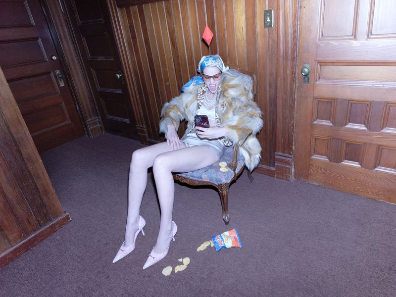 Model seated in armchair holding a phone wearing fur coat and headscarf with floating red diamond overhead