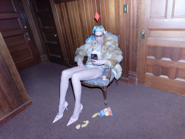 Model seated in armchair holding a phone wearing fur coat and headscarf with floating red diamond overhead
