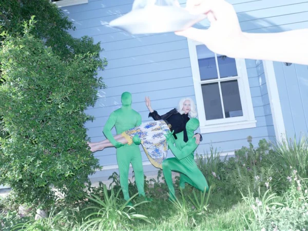 Model Abby Champion held horizontally by two figures in green bodysuits in front of a blue house for Rouge Fashionbook.