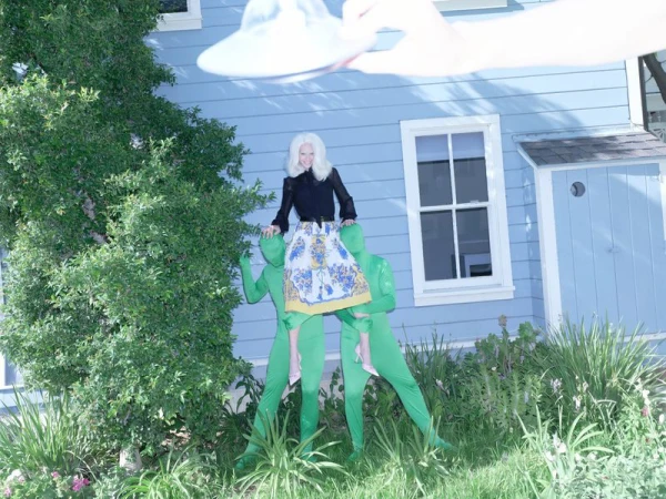 Model perched on two people in green bodysuits before a blue house with a hand holding a hat above