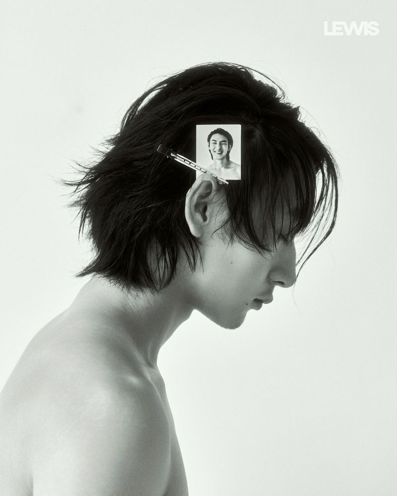Monochrome portrait of a young man with dark medium-length hair holding a small photo clipped to his hair against a white background.