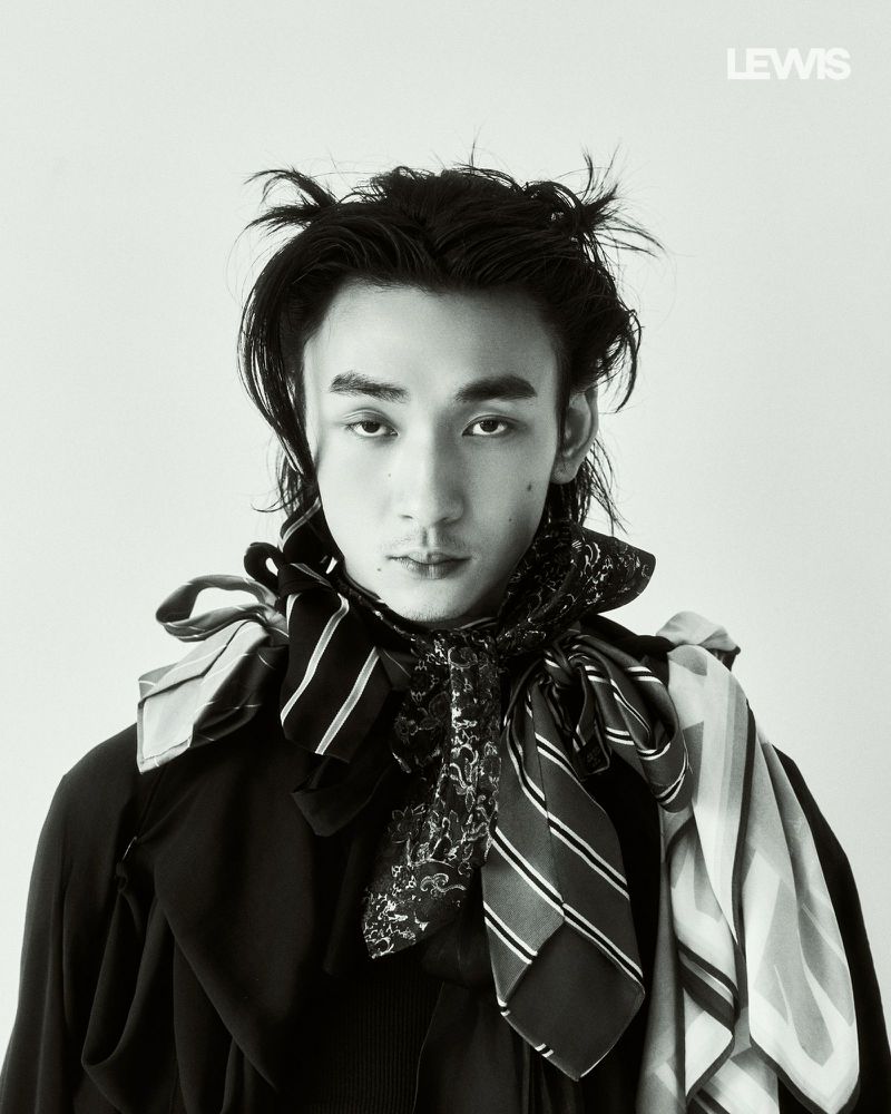 Portrait of a man with styled dark hair wearing multiple patterned scarves and a dark top against a plain light background