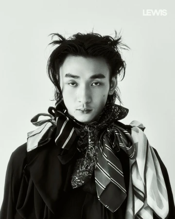 Portrait of a man with styled dark hair wearing multiple patterned scarves and a dark top against a plain light background