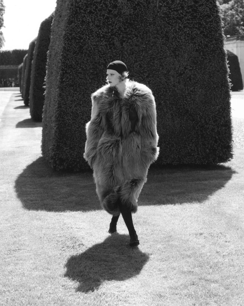 Model in a dark cap and voluminous fur coat walking past manicured hedges for V Magazine