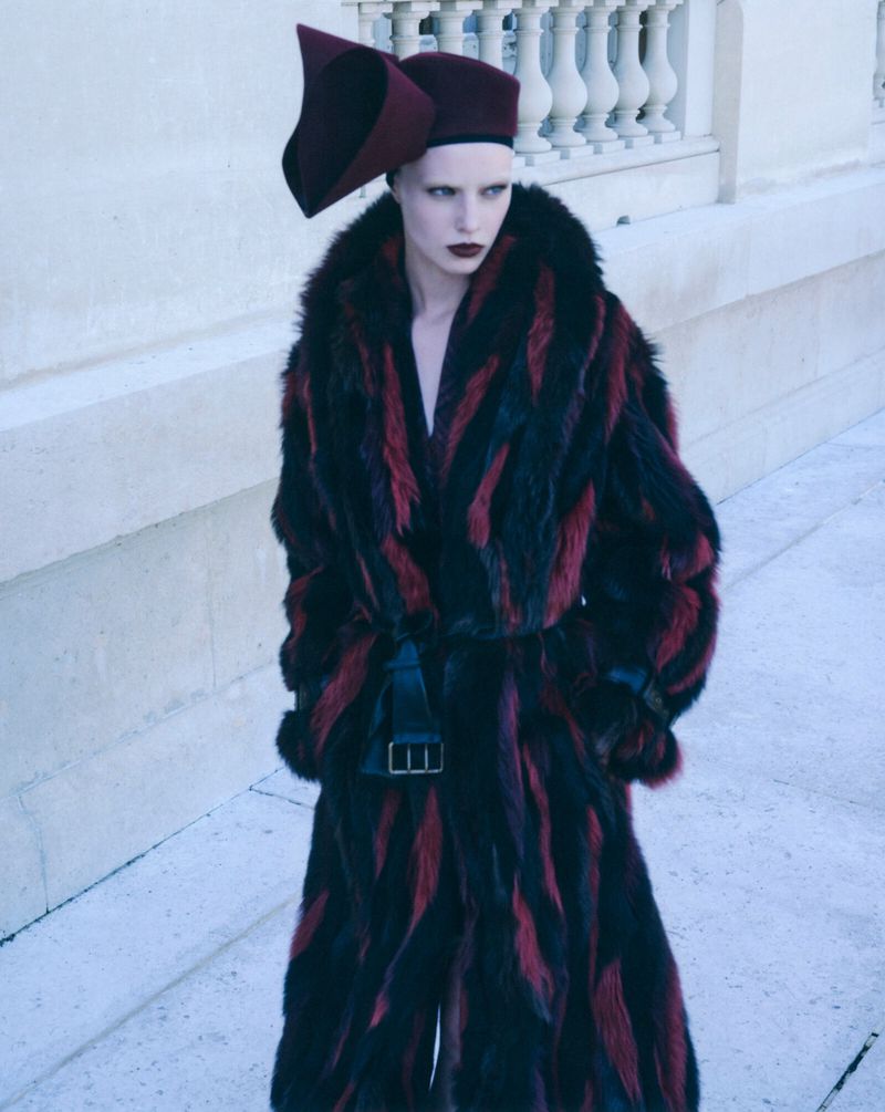 Model wearing a black and red patterned faux fur coat and structured burgundy hat outdoors against a building exterior.
