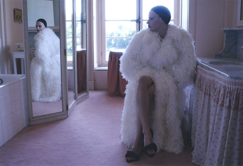 Model seated in a fluffy white textured coat inside a pink room with a mirror reflecting her profile.
