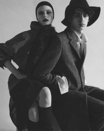 Two models in dark layered coats and hats posing against a plain background in a dramatic fashion studio portrait.
