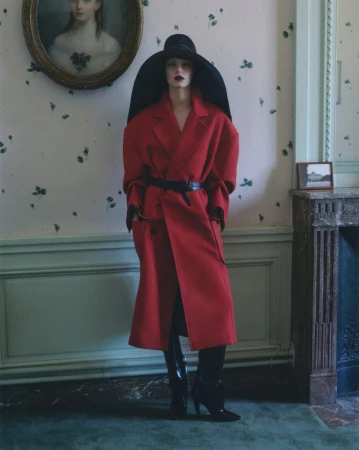 Model in a long red coat and large black hat standing against floral wallpaper for V Magazine editorial