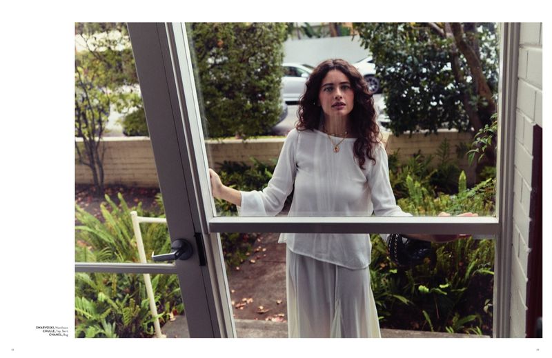 Woman with long dark curly hair wearing a white top standing inside looking out a glass door into a green garden.