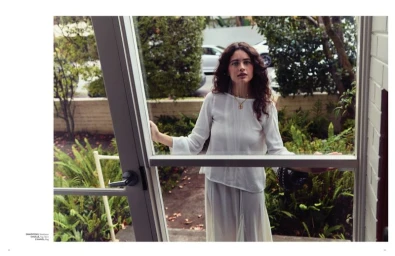 Woman with long dark curly hair wearing a white top standing inside looking out a glass door into a green garden.