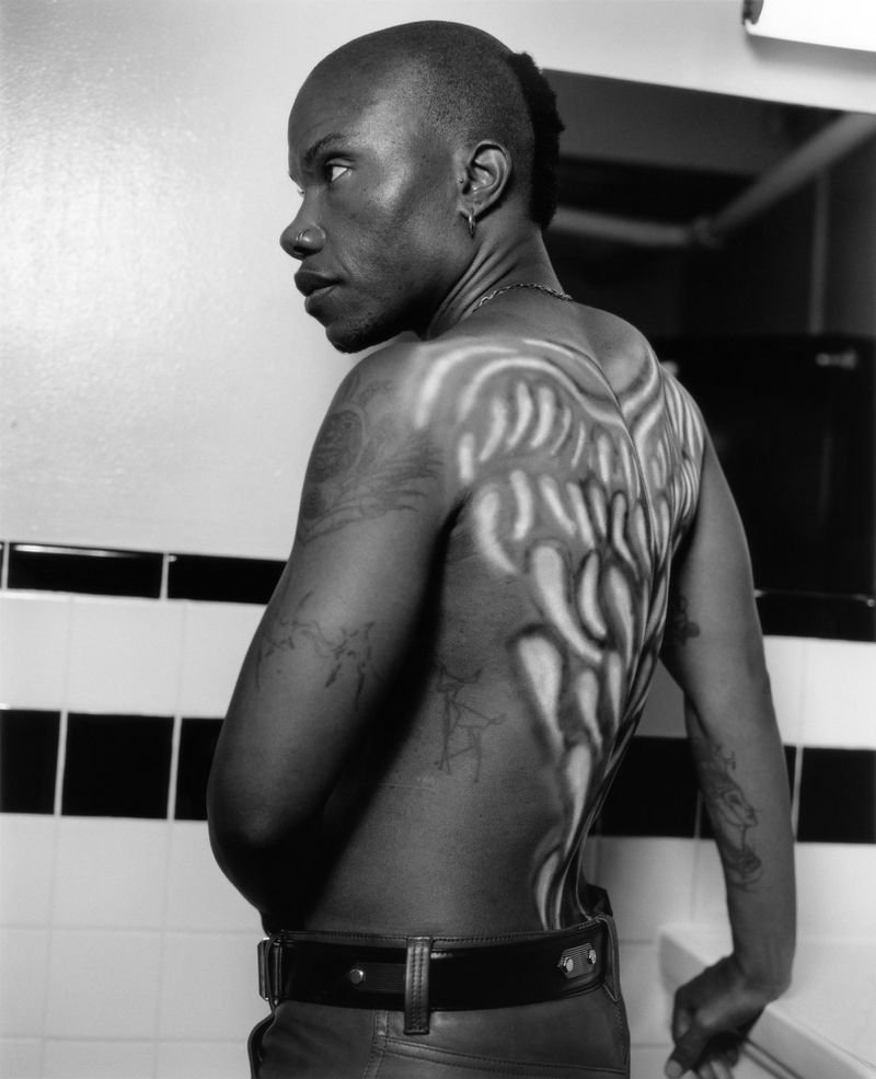 Black man with airbrushed angel wings on his bare back looking over his shoulder in a tiled bathroom setting.