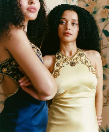 Two women with dark curly hair wearing embellished halter dresses posing against a patterned wallpaper background.