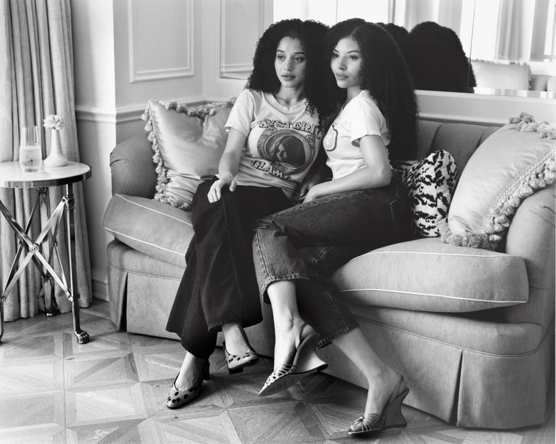 Two women seated together on a light fabric sofa in a room with parquet flooring and curtains