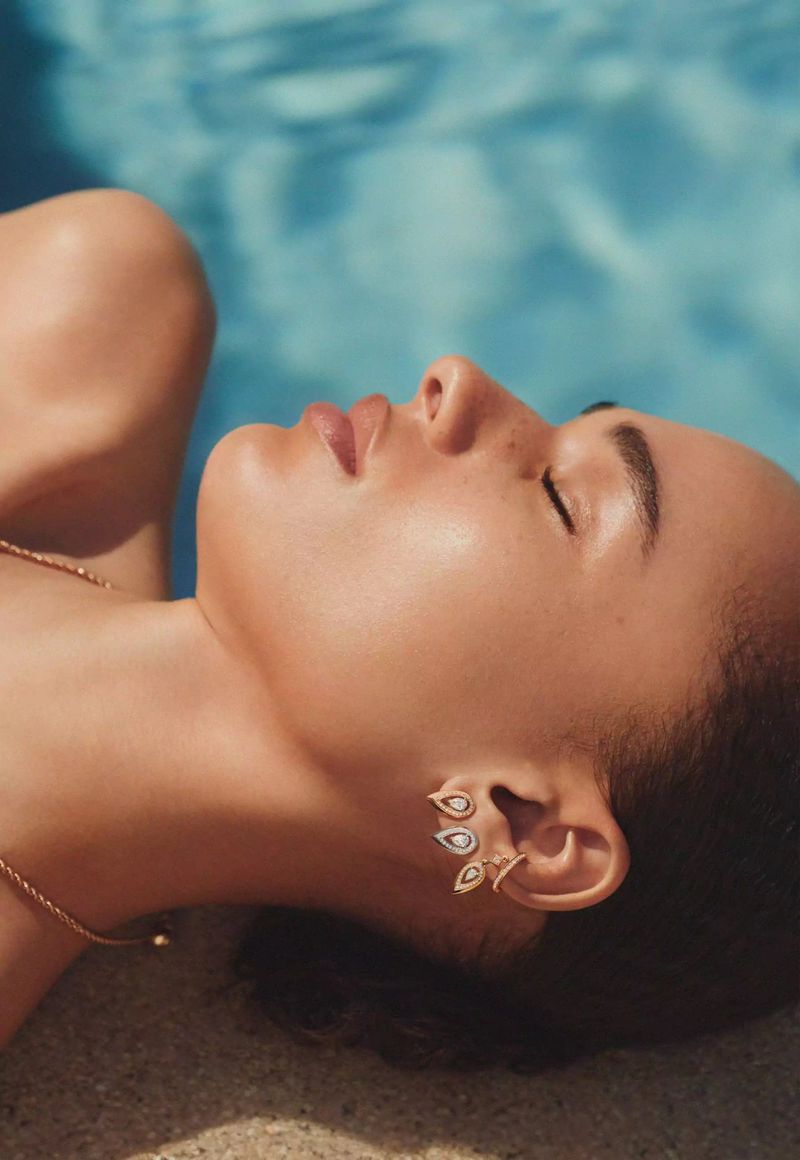 Close-up of a woman with jewelry lying outdoors in sunlight, with a pool and sand visible in the background on a bright day.