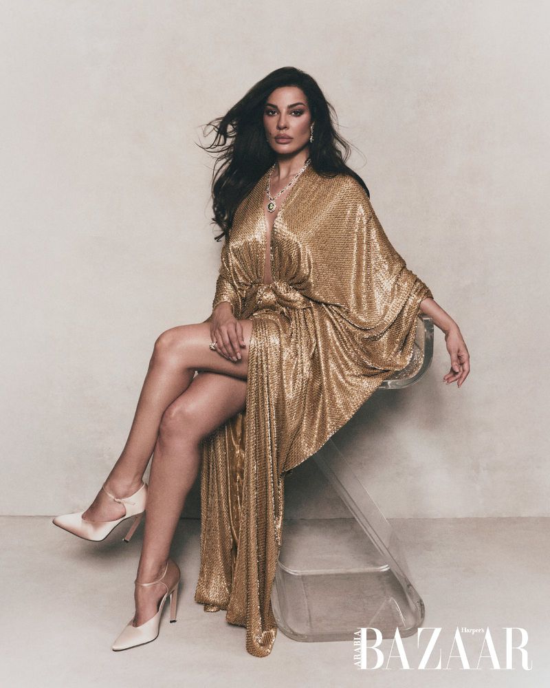Woman in a sparkling gold dress seated on a transparent acrylic stool against a neutral textured studio background.